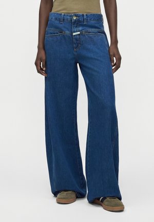 Personne portant un jean bleu taille haute à jambes larges et des baskets vert olive, debout devant un fond clair uni.