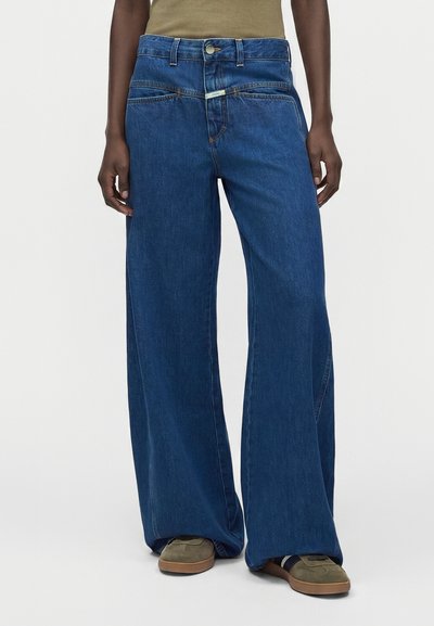 Personne portant un jean bleu taille haute à jambes larges et des baskets vert olive, debout devant un fond clair uni.