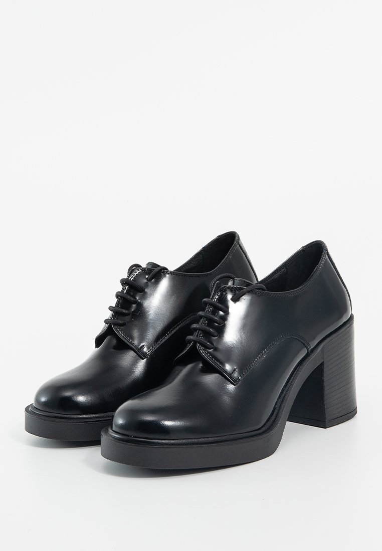 Une paire de chaussures en cuir verni noir à lacets avec des orteils ronds et des talons épais sur fond blanc.
