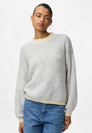 Suéter de color beige claro con un patrón de diamantes texturizado, con cuello redondo y mangas holgadas, combinado con jeans azules.