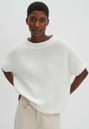 Donna sicura di sé con capelli corti che indossa una maglia bianca larga a maglia e pantaloni beige, in piedi con le mani sui fianchi contro uno sfondo chiaro.