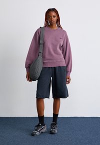 Woman with red braids wearing a purple sweatshirt, black knee-length shorts, black socks, sneakers, and carrying a checkered shoulder bag.