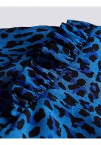 Sininen eläinkuosinen kangas mustilla täplillä, jossa on rypytetty röyhelöyksityiskohta reunassa. Sileä pinta ja elävä värikontrasti.