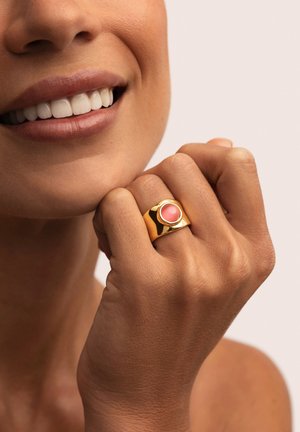 Anillo de oro con una piedra rosa redondeada, con una textura suave y una banda ancha. La mano está colocada cerca de la cara, mostrando un tono de piel natural.