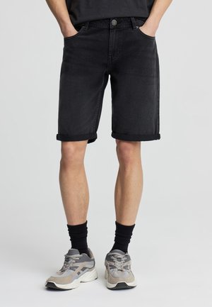 Persona che indossa pantaloncini di jeans neri con orlo arrotolato, calzini neri e scarpe da ginnastica grigie, in piedi con le mani in tasca su sfondo chiaro.