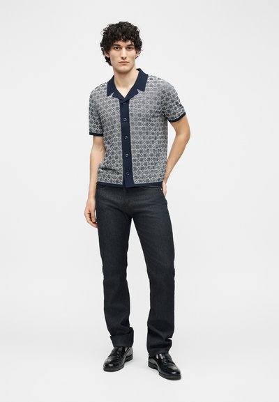 Jeune homme aux cheveux bouclés portant une chemise à manches courtes à motifs, un jean foncé et des chaussures en cuir noir, debout avec une main dans la poche.