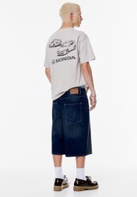 Lys grå T-shirt med sorte bilgrafikker og "HONDA" tekst; parret med mørke denimshorts med store lommer og et tanmærke.