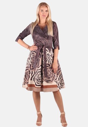 Frau steht mit der Hand in der Hüfte und trägt ein knielanges braun-beiges Blumenmusterkleid mit gebundener Taille und beigen hochhackigen Sandalen.