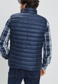 Chaleco acolchado de color azul marino con acolchado horizontal, cuello alto y cierre de cremallera. Lleva encima una camisa de cuadros azul y blanca de manga larga.