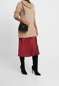 Suéter de punto beige con cuello cisne, combinado con una falda de satén burdeos, botas de tacón de aguja negras por encima de la rodilla y un bolso negro texturizado.