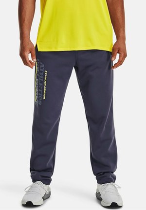 Pantaloni sportivi blu navy con fascia elastica in vita, tasche laterali e grafica prominente "Athletics" in giallo sulla gamba sinistra. Abbinati a scarpe da ginnastica bianche.