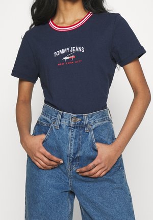 Vrouw in een marineblauw Tommy Jeans t-shirt met een rode en witte kraag, gestoken in hoge blauwe denim jeans, met handen in de zakken.