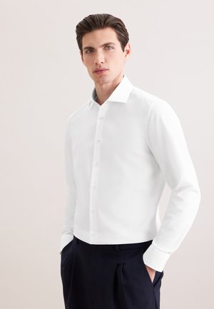Homme portant une chemise blanche boutonnée rentrée dans un pantalon noir, mains dans les poches, sur un fond clair uni.
