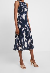 Abito midi tie-dye blu navy e crema con corpetto aderente, design senza maniche e orlo svasato. Abbinato a sandali con tacco a fascia beige.
