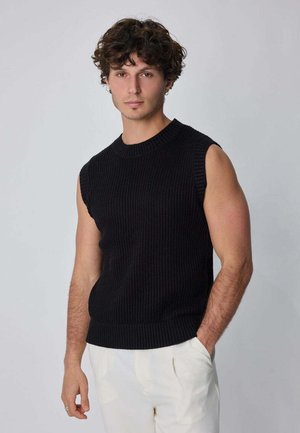 Jeune homme aux cheveux bouclés portant un pull sans manches en tricot noir et un pantalon blanc, debout avec une main dans la poche devant un fond gris.