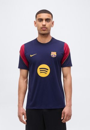 Młody mężczyzna z krótkimi włosami, ubrany w granatowo-czerwoną koszulkę piłkarską FC Barcelona z żółtymi logotypami Nike i Spotify, stoi na jednolitym tle.