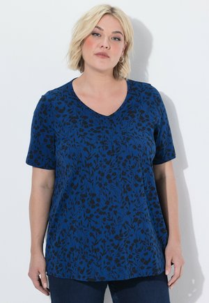 Kvinde med kort blondt hår iført en blå V-halsbluse med sort blomsterprint og mørke bukser mod en hvid baggrund.