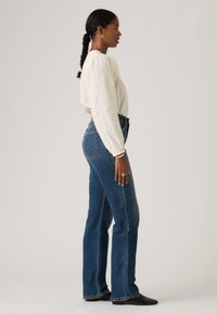 Γυναίκα με πλεξούδες, φορώντας λευκό υφασμάτινο πουκάμισο, τζιν ψηλόμεση μπλε, μαύρα μυτερά παπούτσια και δαχτυλίδι, όρθια σε προφίλ.