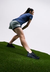 Baskets de sport noires avec des semelles texturées, associées à une chemise bleue et un short gris, présentées sur une surface herbeuse. Accent sur la posture et le mouvement.
