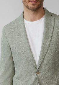 Jasnozielona marynarka o teksturowym wykończeniu z wykładanym kołnierzem, na pojedynczy guzik i kieszenią naszywaną, noszona na białym t-shircie.