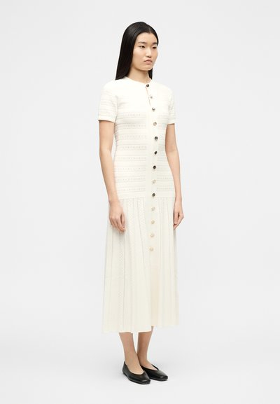 Μια γυναίκα φοράει ένα midi φόρεμα με κοντά μανίκια σε λευκό χρώμα με υφασμένους οριζόντιους ρίγες  και κουμπιά μπροστά, συνδυασμένο με μαύρα μπαλαρίνες.