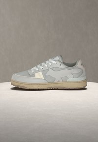Baskets basses gris clair avec lacets blancs, semelle beige et un subtil motif en forme de chien sur le côté, présentées sur une surface claire.