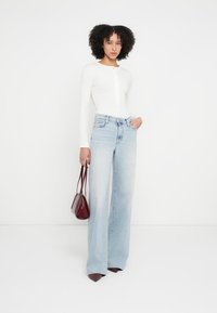 Balta garām piedurknēm topiņa ar pogu detaļām, kopā ar gaiši mazgātām platajām džinsa biksēm. Modelis rokā tur nelielu bordo krāsas somiņu.