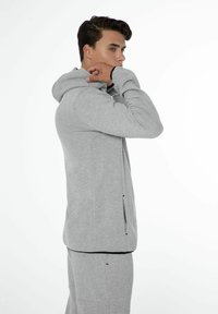 Sweat-shirt à capuche gris avec une fermeture éclair sur le devant, des poches latérales et une coupe décontractée. Texture douce et tissu lisse. Porté avec un pantalon assorti.