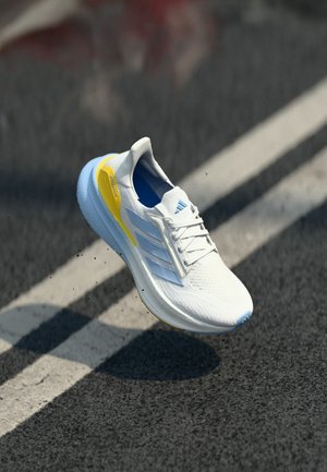 Chaussure de sport blanche avec des accents bleus et jaunes flottant au-dessus d'une route en asphalt avec des lignes blanches, projetant une ombre en dessous.