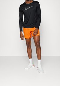 Mann in einem schwarzen Langarmshirt von Nike, leuchtend orangefarbigen Nike-Shorts, weißen Socken und weißen Turnschuhen vor einem schlichten Hintergrund.