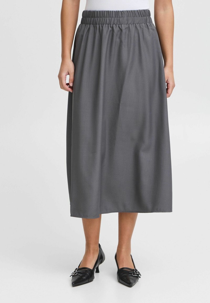 Femme portant une jupe midi grise avec une taille élastique et des chaussures noires à talon bas et bout pointu, debout sur un fond uni.