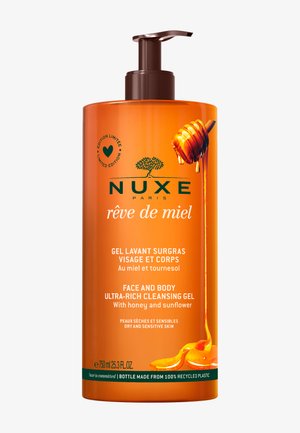 Orange, rundt flaske med pumpe-dispenser. Har et grafisk design af honningdrizzle. Teksten inkluderer produktdetaljer på fransk og engelsk, med sort-hvide accenter.