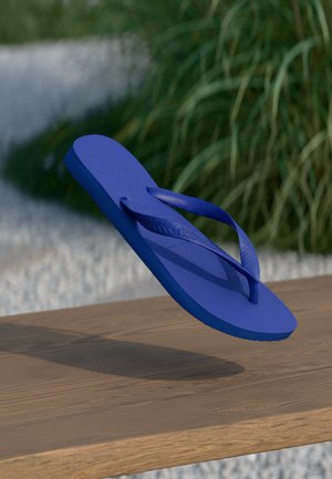 Flip-flop din cauciuc albastru, cu partea superioară texturată, talpă plată și bretele în formă de V, suspendat deasupra unei suprafețe din lemn, cu iarbă în fundal.