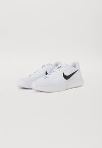 Pár bílých běžeckých tenisek Nike s černým logem swoosh, šněrovací design a texturované gumové podrážky na čistě bílém pozadí.
