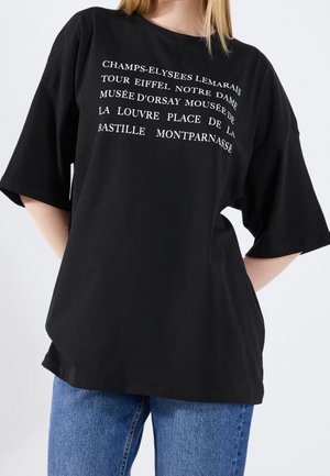 Osoba nosząca czarny oversize'owy t-shirt z białym napisem wymieniającym zabytki Paryża, w połączeniu z niebieskimi dżinsami, ręce za plecami.