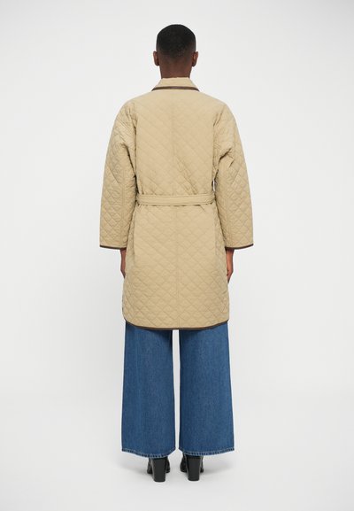 Beige quilted coat with a belted waist, featuring a collar, long sleeves, and brown trim. Paired with wide-leg denim trousers and heeled boots.