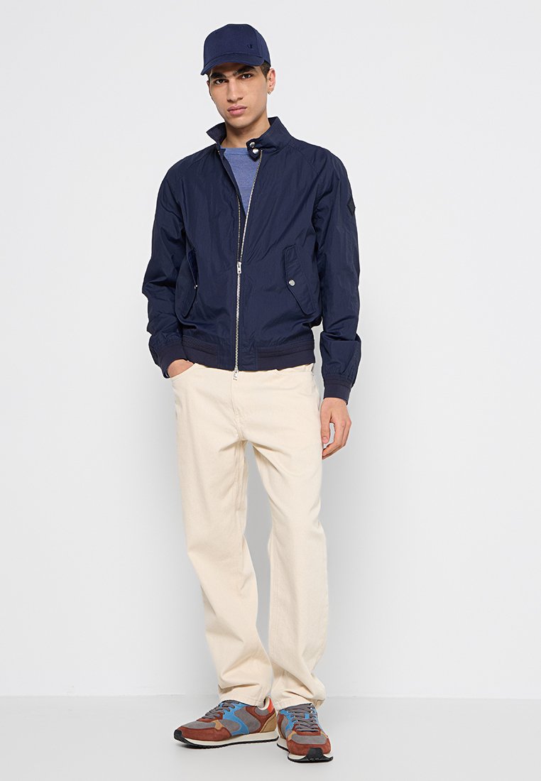 Junger Mann, der eine marineblaue Mütze, eine marineblaue Jacke mit Reißverschluss, ein blaues Hemd, cremefarbene Hosen und mehrfarbige braune und blaue Sneaker trägt, steht vor einem weißen Hintergrund.