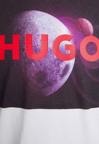 Camiseta gráfica con fondo oscuro y diseño espacial, gran texto "HUGO" en rojo y gráfico de una luna púrpura con planetas.