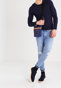 Niebieska torebka na pasku z brązowymi akcentami, noszona z dopasowaną czarną koszulą z długim rękawem, jasnymi, lekko znoszonymi dżinsami oraz ciemnymi zamszowymi butami.