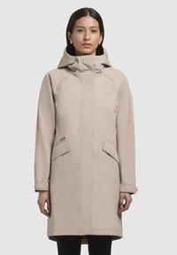 Parka beige imperméable avec capuche, dotée de poches latérales, d'une silhouette légèrement ajustée et d'une texture lisse. Design simple et fonctionnel.