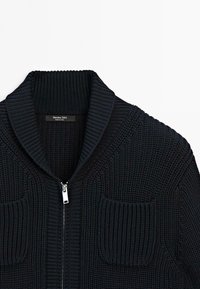 Close-up of a black chunky knit zip-up sweater with ribbed shawl collar and two front patch pockets.