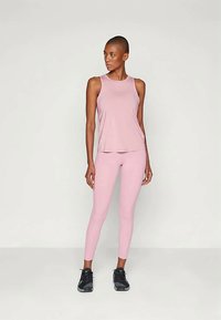 Moteris, vilkinti be Rankovių rožinį sportinį viršų ir atitinkamas legginus, stovi su kojomis pečių plotyje, žiūri į dešinę, avėdama juodus sportinius batelius.