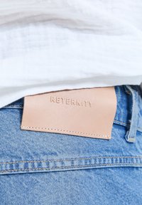 Parche de cuero beige con la palabra "RETERNITY" en relieve en la pretina de unos vaqueros de mezclilla azul bajo una camisa blanca.
