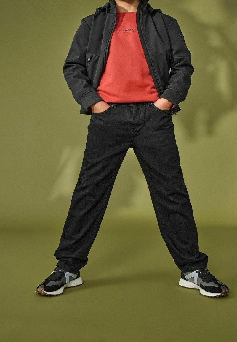 Personne debout, mains dans les poches, portant une veste noire, une chemise rouge, un pantalon noir et des baskets noir et blanc sur fond vert.