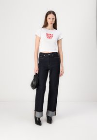 T-shirt cropped branco com texto vermelho; jeans de perna larga em denim escuro com bainhas azuis dobra; bolsa preta; botas pretas de bico fino.