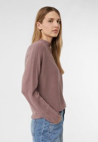 Helllila Pullover mit hohem Kragen und gerippten Seitendetails. Gestrickte Textur mit langen Ärmeln und lässiger Passform. Kombiniert mit hellblauen Jeans.