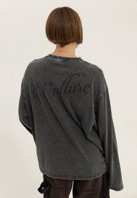 Yläripsinen hiilenharmaa collegepaita, jossa on pehmeä tekstuuri. "Kulttuuri" painettuna tummalla fontilla selkäpuolella. Pitkät hihat ja roikkuvat olkapäät.