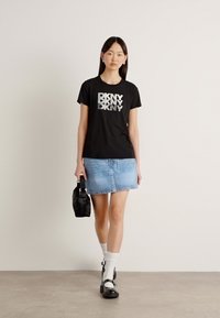 T-shirt di cotone nera con un logo grafico argentato "DKNY", abbinata a una gonna di jeans chiari, calze bianche e scarpe nere. Accessorio borsa semplice.