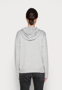 Kobieta z ciemnymi włosami związanymi w kok, ubrana w jasnoszary hoodie i czarne dżinsy, stojąca przodem do jednolitego białego tła.