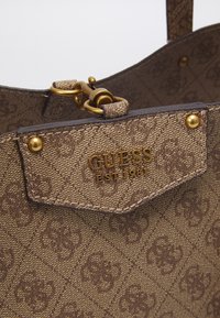 Hnědá kabelka se vzorem, kterou charakterizuje strukturovaný design, zlaté kovové detaily a texturovaná látka s výrazným logem značky.